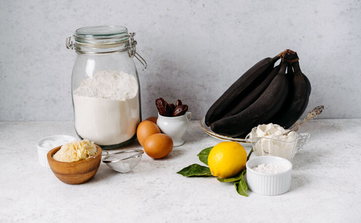 Ingredients for a healthy birthday cake recipe, including overripe bananas, flour, eggs, dates, lemon and cream cheese.